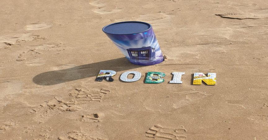 Rob's bespoke scatter tube with Tardis print placed on the sandy beach with letters spelling 'Robin' beside it