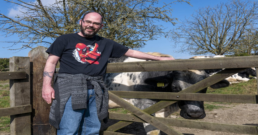 Rob standing in front of a wooden gate in the countryside with a cow behind him